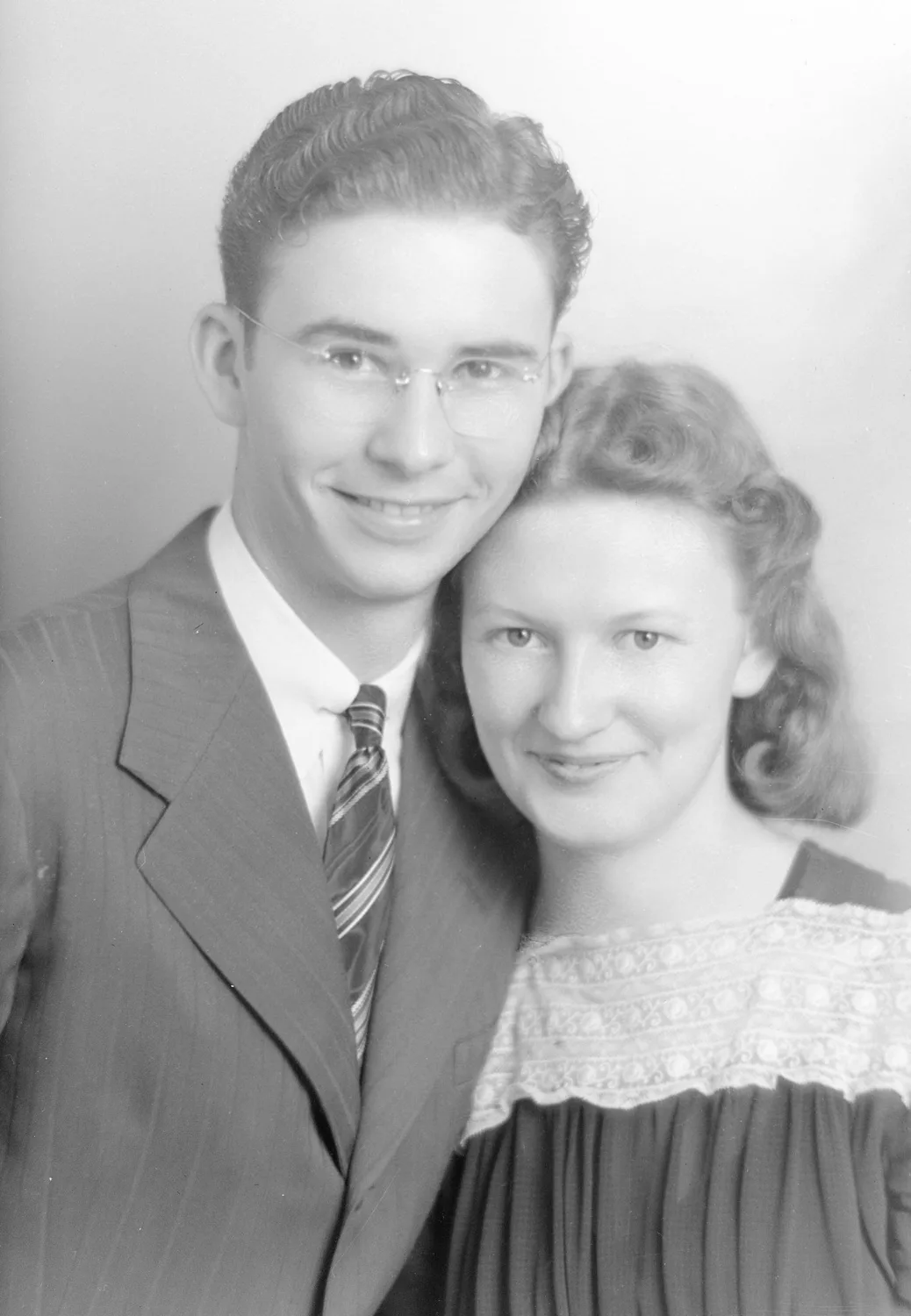 Portrait of Rayford and Oneta Anderson. Rayford is on the left with medium toned slicked back hair and round wire glasses and wears a pin stripe stuit and tie. He puts his arm around Oneta, who has medium toned short curled hair and wears a dark blouse with ruffles and a band of lace across the chest.
