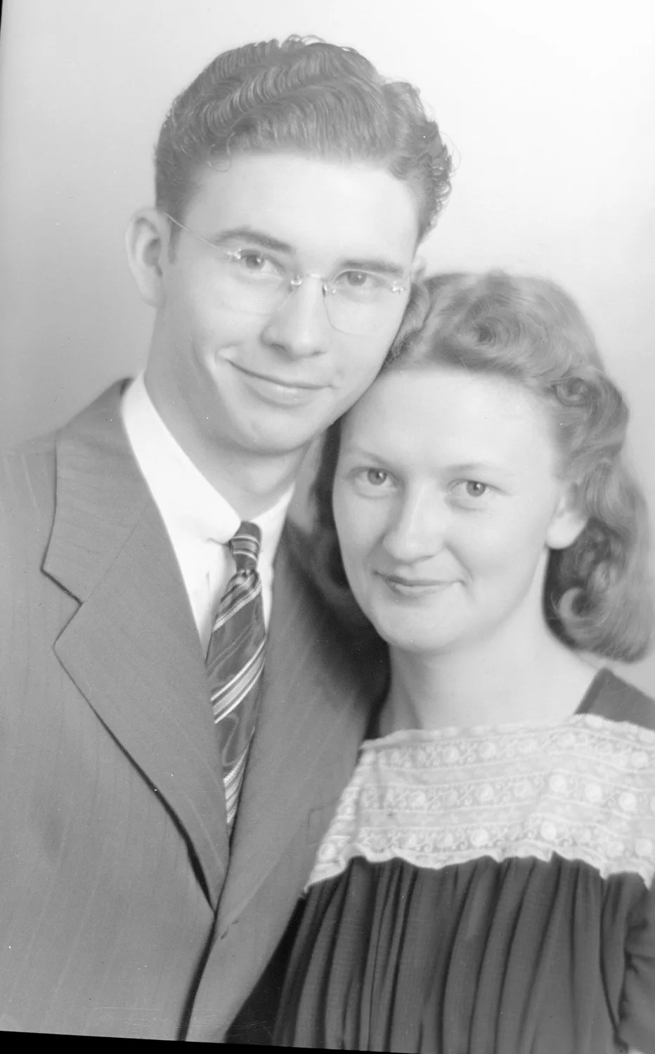 Portrait of Rayford and Oneta Anderson. Rayford is on the left with medium toned slicked back hair and round wire glasses and wears a pin stripe stuit and tie. He puts his arm around Oneta, who has medium toned short curled hair and wears a dark blouse with ruffles and a band of lace across the chest.