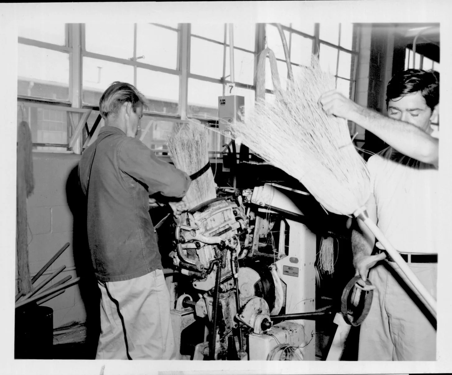 Stitching machine operator stitches one broom as worker prepares another note band in inmate's hand, right used during stitching.